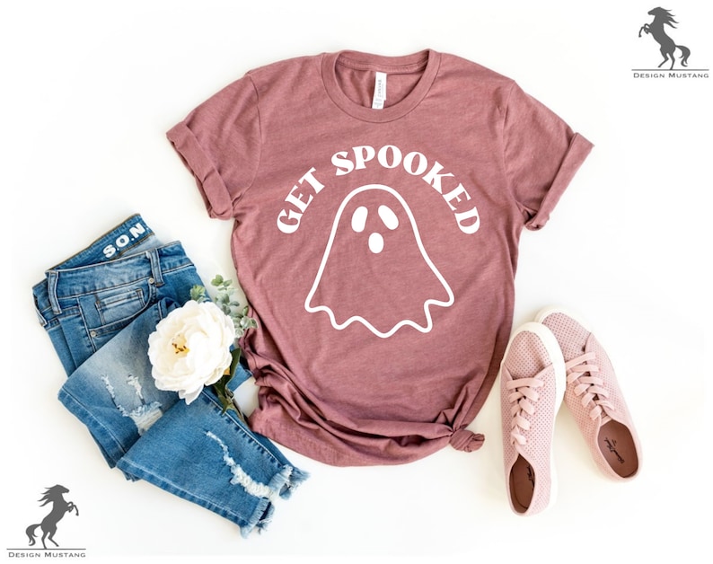 K&ouml;nnte beinhalten: Ein mauvefarbenes T-Shirt mit einer wei&szlig;en Geistergrafik und dem Text "GET SPOOKED". Das Shirt liegt auf einer wei&szlig;en Oberfl&auml;che mit einem Paar rosa Sneakers und einer blauen Jeans.