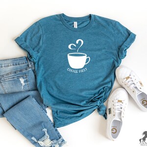 May include: A teal blue t-shirt with a white graphic of a coffee cup with steam and the text "Coffee First". The shirt is laid out on a white surface with a pair of blue jeans and white trainers.