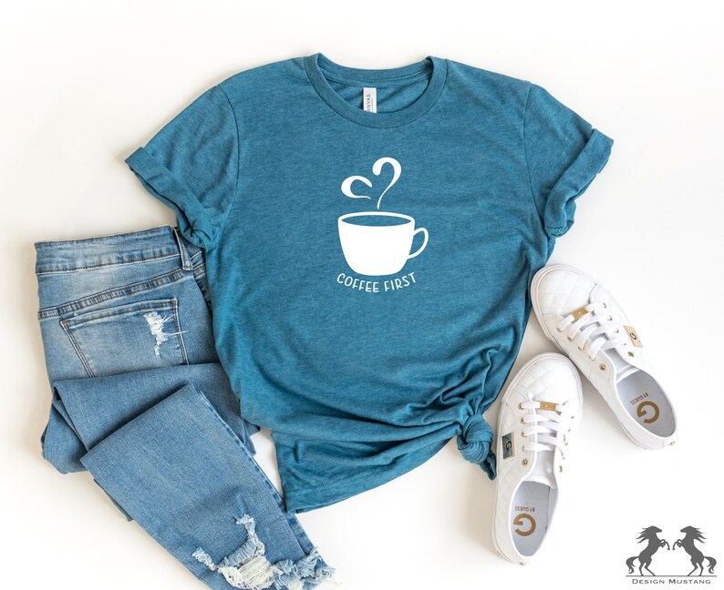 May include: A teal blue t-shirt with a white graphic of a coffee cup with steam and the text "Coffee First". The shirt is laid out on a white surface with a pair of blue jeans and white trainers.