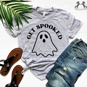 K&ouml;nnte beinhalten: Ein graues T-Shirt mit einer schwarzen Geistergrafik und dem Text "GET SPOOKED" darauf gedruckt. Das Shirt liegt auf einer wei&szlig;en Oberfl&auml;che mit einem Paar braunen Sandalen und einer blauen Jeans.