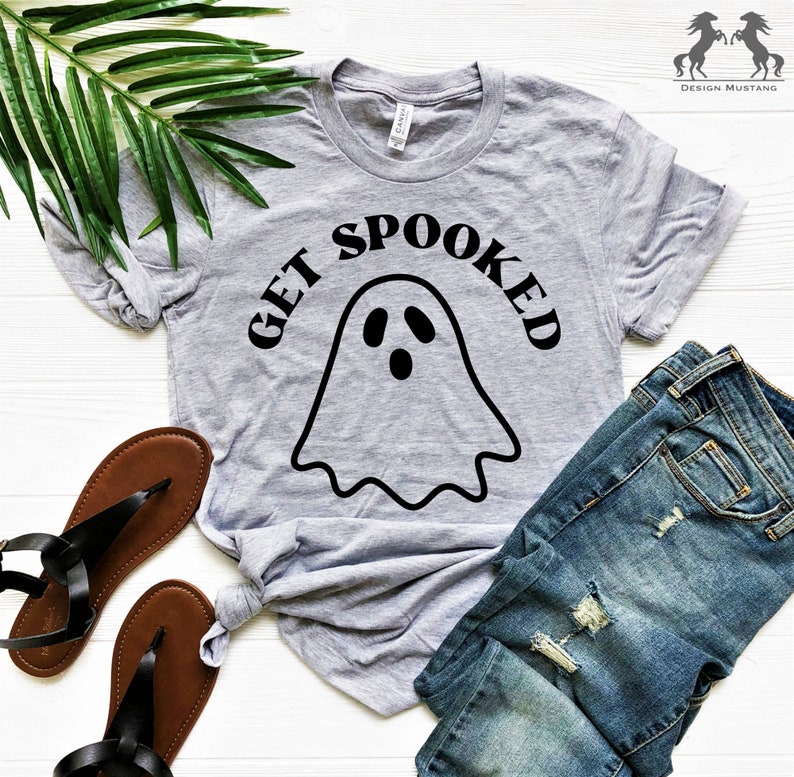 K&ouml;nnte beinhalten: Ein graues T-Shirt mit einer schwarzen Geistergrafik und dem Text "GET SPOOKED" darauf gedruckt. Das Shirt liegt auf einer wei&szlig;en Oberfl&auml;che mit einem Paar braunen Sandalen und einer blauen Jeans.