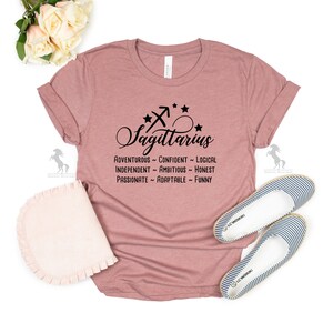 May include: A dusty rose t-shirt with a black Sagittarius zodiac symbol and the text "Sagittarius" in a cursive font. The text "Adventurous - Confident - Logical - Independent - Ambitious - Honest - Passionate - Adaptable - Funny" is printed below the symbol.