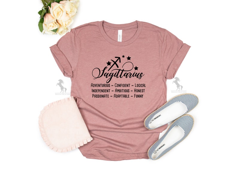 May include: A dusty rose t-shirt with a black Sagittarius zodiac symbol and the text "Sagittarius" in a cursive font. The text "Adventurous - Confident - Logical - Independent - Ambitious - Honest - Passionate - Adaptable - Funny" is printed below the symbol.