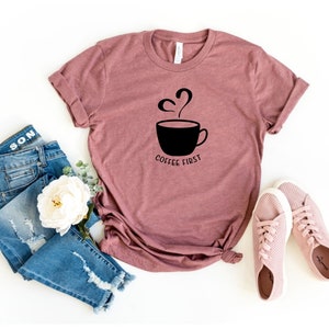 May include: A mauve t-shirt with a black graphic of a coffee cup with steam rising from it and the text "Coffee First". The t-shirt is laid flat on a white surface with a pair of pink trainers, a white flower, and a pair of blue jeans.