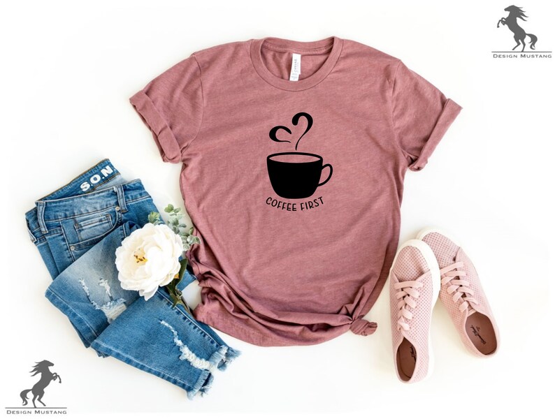 May include: A mauve t-shirt with a black graphic of a coffee cup with steam rising from it and the text "Coffee First". The t-shirt is laid flat on a white surface with a pair of pink trainers, a white flower, and a pair of blue jeans.