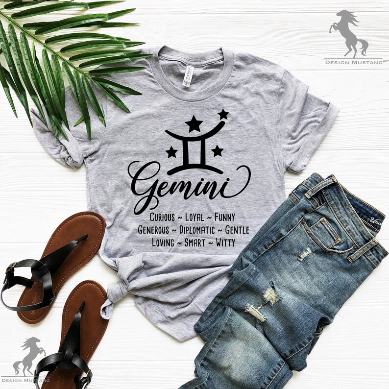 Op de afbeelding: Een grijs T-shirt met een zwart Tweelingen-sterrenbeeld en de tekst "Tweelingen" in een gestileerd lettertype. De tekst "Nieuwsgierig ~ Loyal ~ Grappig ~ Grootmoedig ~ Diplomatiek ~ Lief ~ Liefdevol ~ Slim ~ Vlot" staat onder het symbool.