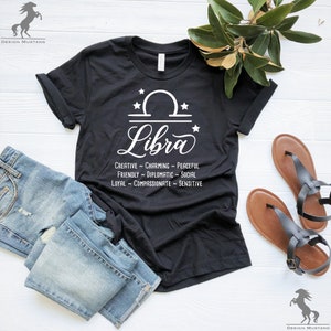 Pu&ograve; includere: Maglietta nera con un segno zodiacale Bilancia bianco e il testo "Libra" in un font bianco corsivo. Il testo sotto il segno recita "Creative ~ Charming ~ Peaceful ~ Friendly ~ Diplomatic ~ Social ~ Loyal ~ Compassionate ~ Sensitive".