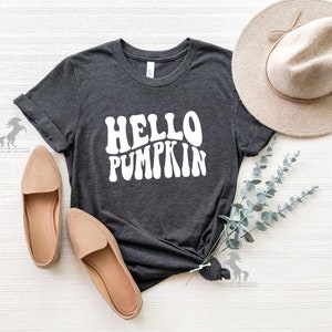 May include: A dark grey t-shirt with the text "HELLO PUMPKIN" in white, stylized in a retro font. The shirt is laid flat on a white surface with a brown hat and green foliage.
