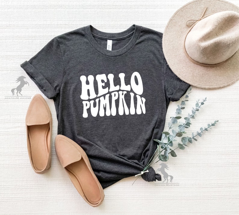May include: A dark grey t-shirt with the text "HELLO PUMPKIN" in white, stylized in a retro font. The shirt is laid flat on a white surface with a brown hat and green foliage.