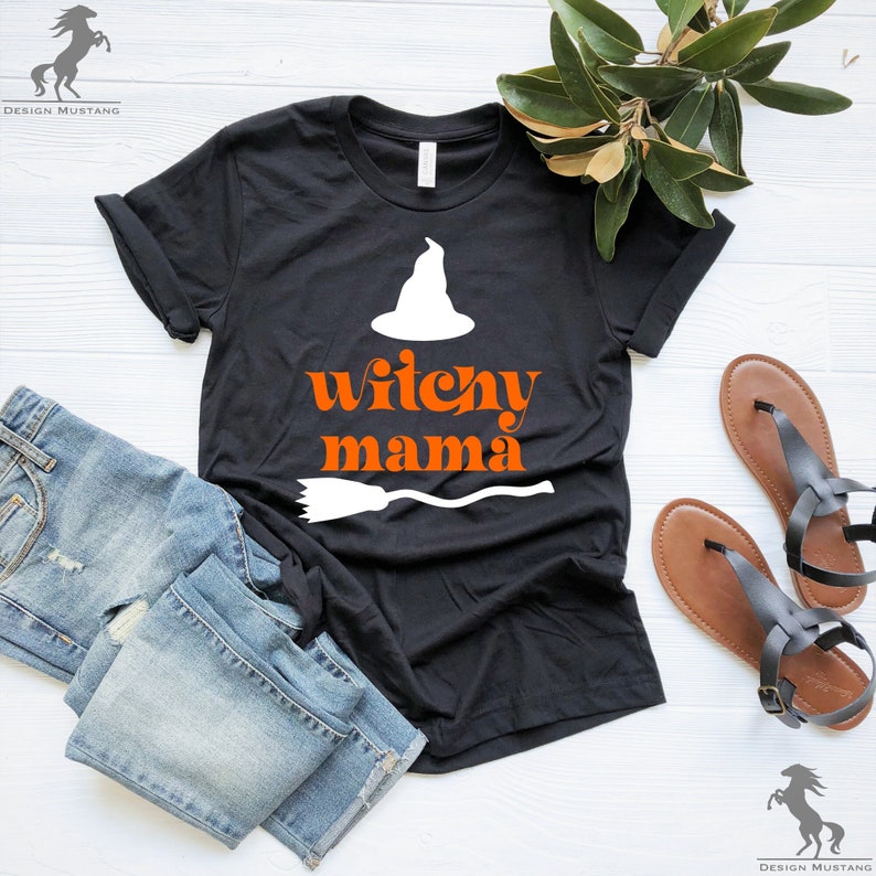 May include: Black t-shirt with a white witch hat and a broom graphic. The text "witchy mama" is printed in orange on the shirt.