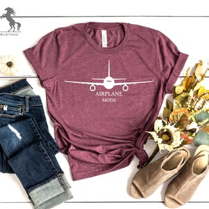 May include: A maroon t-shirt with a white airplane graphic and the text "AIRPLANE MODE". The shirt is laid out on a white wooden surface with a pair of blue jeans, a small white pumpkin, and a bouquet of dried flowers.