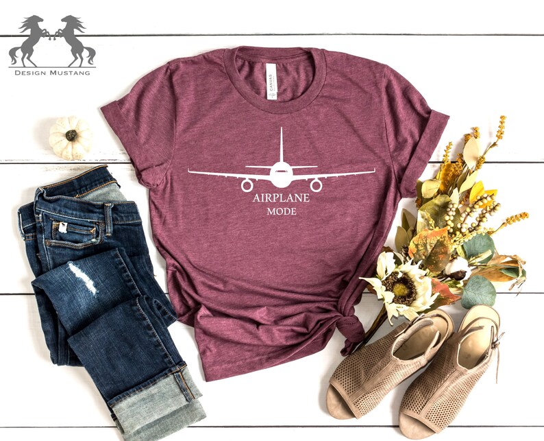 May include: A maroon t-shirt with a white airplane graphic and the text "AIRPLANE MODE". The shirt is laid out on a white wooden surface with a pair of blue jeans, a small white pumpkin, and a bouquet of dried flowers.