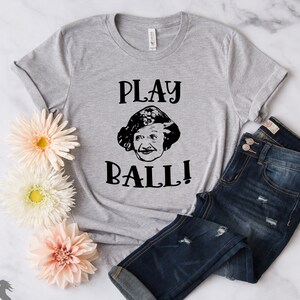 Peut inclure: Un t-shirt gris avec un motif noir repr&eacute;sentant le visage d'une femme et le texte "PLAY BALL!"