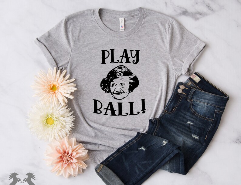 Peut inclure: Un t-shirt gris avec un motif noir repr&eacute;sentant le visage d'une femme et le texte "PLAY BALL!"