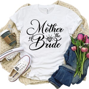 May include: White t-shirt with black text that reads "Mother of the Bride" with a diamond graphic. The shirt is lying on a tan knitted blanket with a pair of white sneakers, a coffee cup, and a bouquet of pink tulips and greenery.