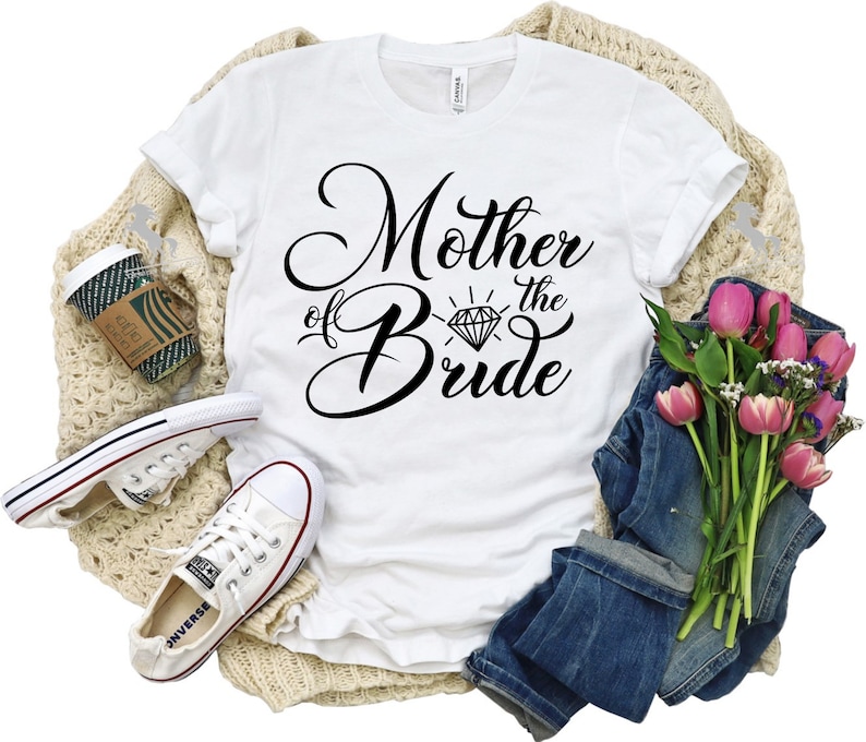 May include: White t-shirt with black text that reads "Mother of the Bride" with a diamond graphic. The shirt is lying on a tan knitted blanket with a pair of white sneakers, a coffee cup, and a bouquet of pink tulips and greenery.