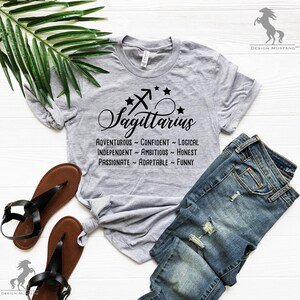 May include: Gray t-shirt with a black Sagittarius zodiac sign and the words "Sagittarius", "Adventurous", "Confident", "Logical", "Independent", "Ambitious", "Honest", "Passionate", "Adaptable", and "Funny".