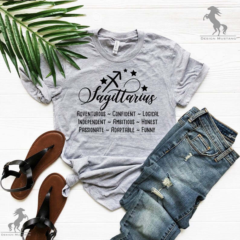 May include: Gray t-shirt with a black Sagittarius zodiac sign and the words "Sagittarius", "Adventurous", "Confident", "Logical", "Independent", "Ambitious", "Honest", "Passionate", "Adaptable", and "Funny".