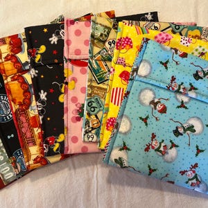 May include: A collection of ten fabric lunch bags with various patterns. The bags are made of cotton and have a variety of designs, including Mickey Mouse, Route 66, and snowmen. The bags are all folded and stacked on top of each other.