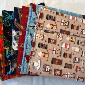 May include: A stack of eight colorful fabric squares with different patterns. The top square features a brown background with a repeating pattern of coffee cups and coffee beans. The other squares feature various patterns, including butterflies, sunflowers, and a red cardinal.