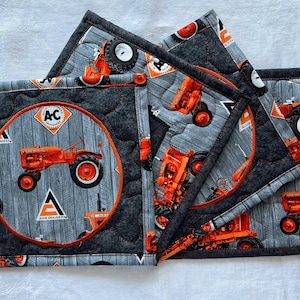 May include: Set of quilted fabric hot pads featuring a gray wood grain pattern with orange tractors. One pad has a large orange circle with a tractor and the text "AC". The pads are edged with black fabric.