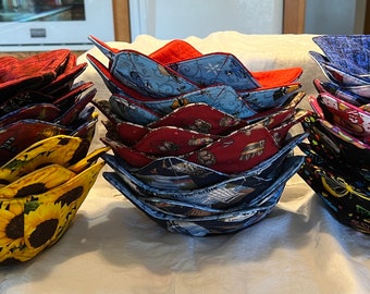 May include: Four stacks of fabric bowl cozies in various patterns. The stacks are arranged from left to right: yellow sunflowers, blue and red floral, red and white floral, and black with colorful cartoon characters.