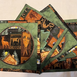 May include: Set of four green and brown fabric coasters with a woodland theme. The coasters feature images of deer, cabins, snowshoes, and trees. The text "Lakeview Cabins" is visible on one of the coasters.