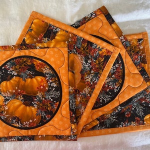May include: Four orange and black quilted pot holders with a pumpkin and fall leaf design. The pot holders have a scalloped edge and a circular design in the center.