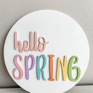 May include: A white circular wooden sign with the words "hello spring" in colorful letters. The letters are pink, purple, blue, orange, yellow, and green.