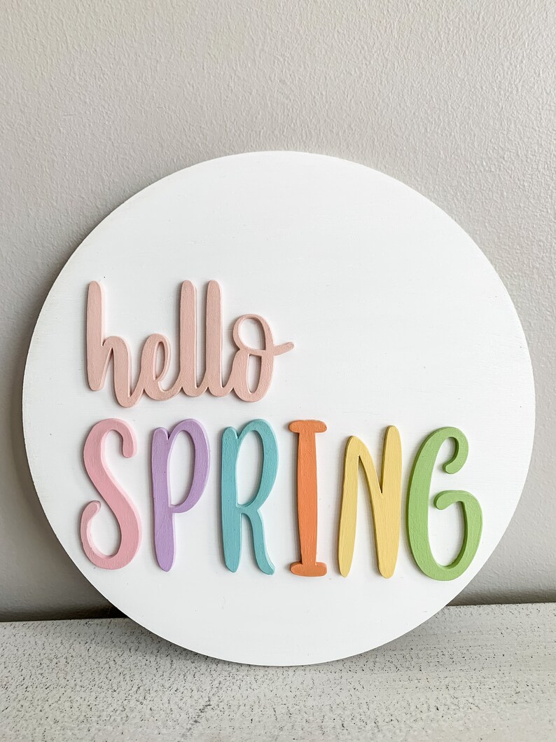 May include: A white circular wooden sign with the words "hello spring" in colorful letters. The letters are pink, purple, blue, orange, yellow, and green.