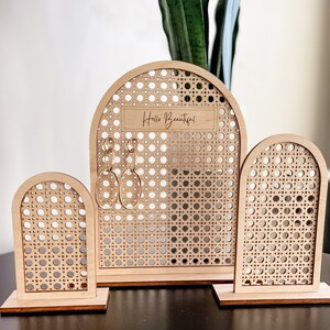May include: A set of three wooden earring displays. The displays have a light brown finish and a woven rattan-style design. The largest display has the text "Hello Beautiful" and holds gold hoop earrings. The other two displays are smaller.