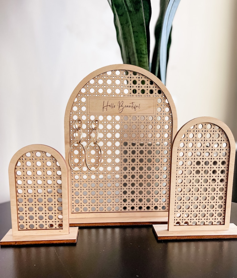 May include: A set of three wooden earring displays. The displays have a light brown finish and a woven rattan-style design. The largest display has the text "Hello Beautiful" and holds gold hoop earrings. The other two displays are smaller.