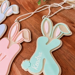 K&ouml;nnte beinhalten: Drei h&ouml;lzerne Hasen-Anh&auml;nger mit rosa, blau und gr&uuml;ner Farbe. Die Anh&auml;nger haben eine Schnur zum Aufh&auml;ngen und die Namen "Kendra", "Avery" und "Lily" sind darauf geschrieben.