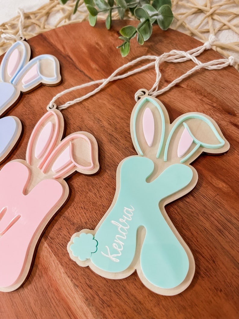 K&ouml;nnte beinhalten: Drei h&ouml;lzerne Hasen-Anh&auml;nger mit rosa, blau und gr&uuml;ner Farbe. Die Anh&auml;nger haben eine Schnur zum Aufh&auml;ngen und die Namen "Kendra", "Avery" und "Lily" sind darauf geschrieben.