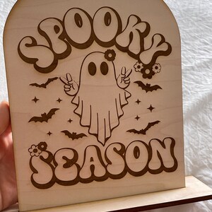 Może przedstawiać: Drewniany znak z duchem noszącym wianek z kwiat&oacute;w i znaki pokoju. Znak głosi "Spooky Season" w retro czcionce.