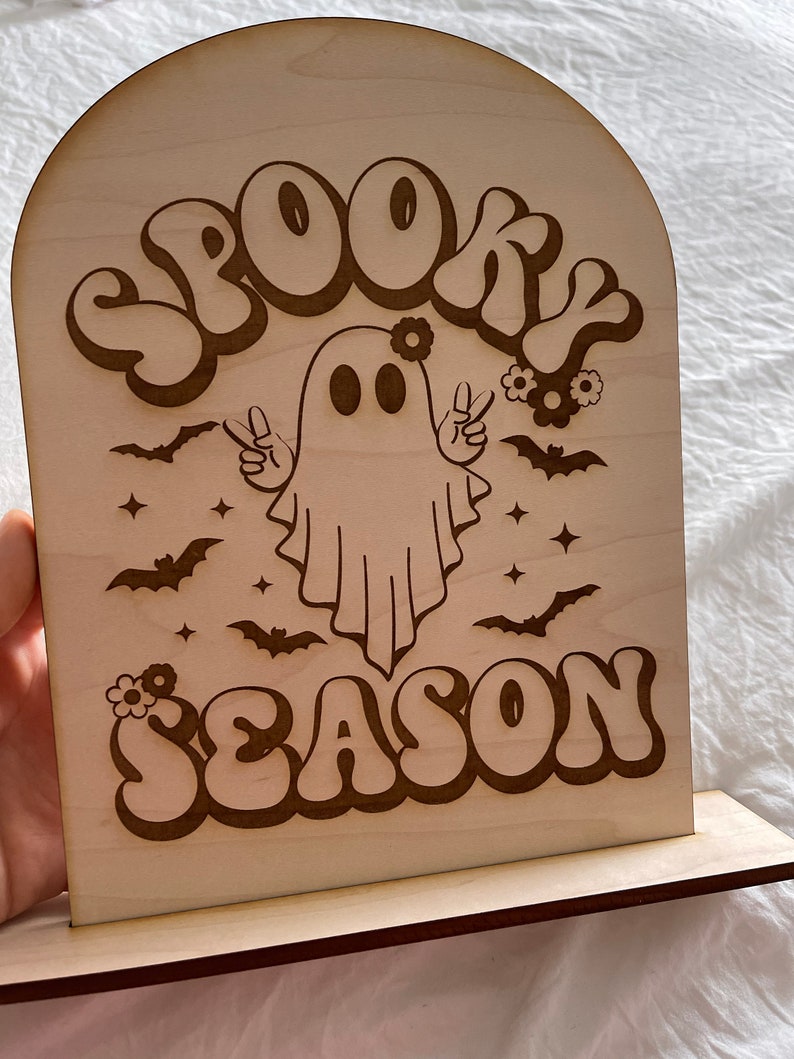 Może przedstawiać: Drewniany znak z duchem noszącym wianek z kwiat&oacute;w i znaki pokoju. Znak głosi "Spooky Season" w retro czcionce.