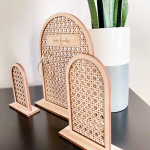 May include: Three wooden earring display stands with a woven rattan design. The largest stand has the text "Hello Beautiful!" engraved on it. The stands are arched and sit on rectangular bases. Gold hoop earrings are displayed on the largest stand.
