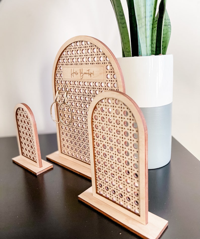 May include: Three wooden earring display stands with a woven rattan design. The largest stand has the text "Hello Beautiful!" engraved on it. The stands are arched and sit on rectangular bases. Gold hoop earrings are displayed on the largest stand.