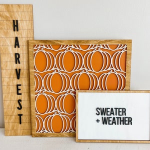 Könnte beinhalten: Drei Holzschilder mit Herbstmotiven und einem Kürbis-Muster. Die Schilder zeigen "HARVEST", "SWEATER + WEATHER" und ein Muster aus weißen Kürbissen auf orangefarbenem Hintergrund.