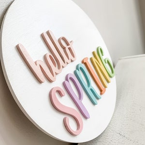 May include: A white round wooden sign with the words "hello spring" written in rainbow colors. The letters are raised and painted in pastel shades of pink, purple, blue, green, and yellow.