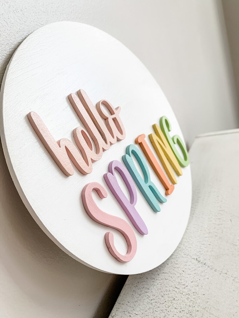 May include: A white round wooden sign with the words "hello spring" written in rainbow colors. The letters are raised and painted in pastel shades of pink, purple, blue, green, and yellow.