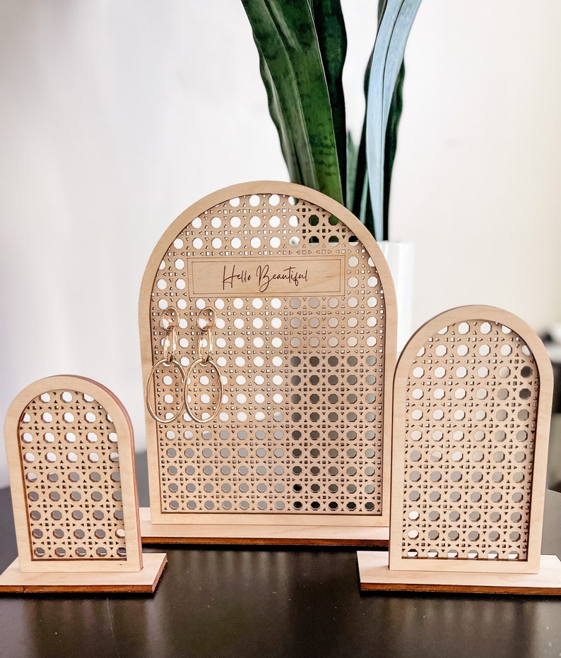 May include: A set of three wooden earring displays. The displays have a light wood finish and a woven cane pattern. The largest display has the words "Hello Beautiful" engraved on it. Gold earrings hang from the center display.