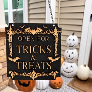 Könnte beinhalten: Ein quadratisches, schwarzes Hängeschild mit einem lasergeschnittenen Holzrahmen, der Fledermäuse und florale Ornamente zeigt. Das Schild trägt den Text "OPEN FOR TRICKS & TREATS" in hellen, erhabenen Buchstaben und hängt an einer Kette.