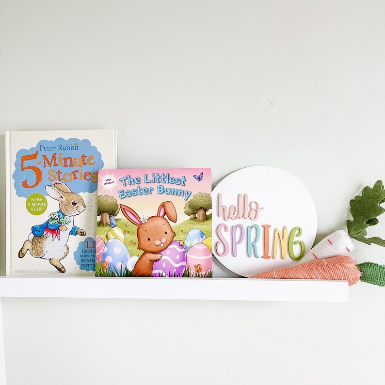 May include: A white shelf with three items on it. The items are a book titled "5 Minute Stories" featuring a rabbit, a book titled "The Littlest Easter Bunny" featuring a bunny, and a round white sign with the text "hello SPRING" in rainbow colors.