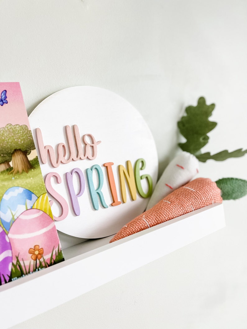 May include: A white round sign with the words "hello spring" in rainbow colors. The sign is on a white shelf with a stuffed carrot and a book with Easter eggs.