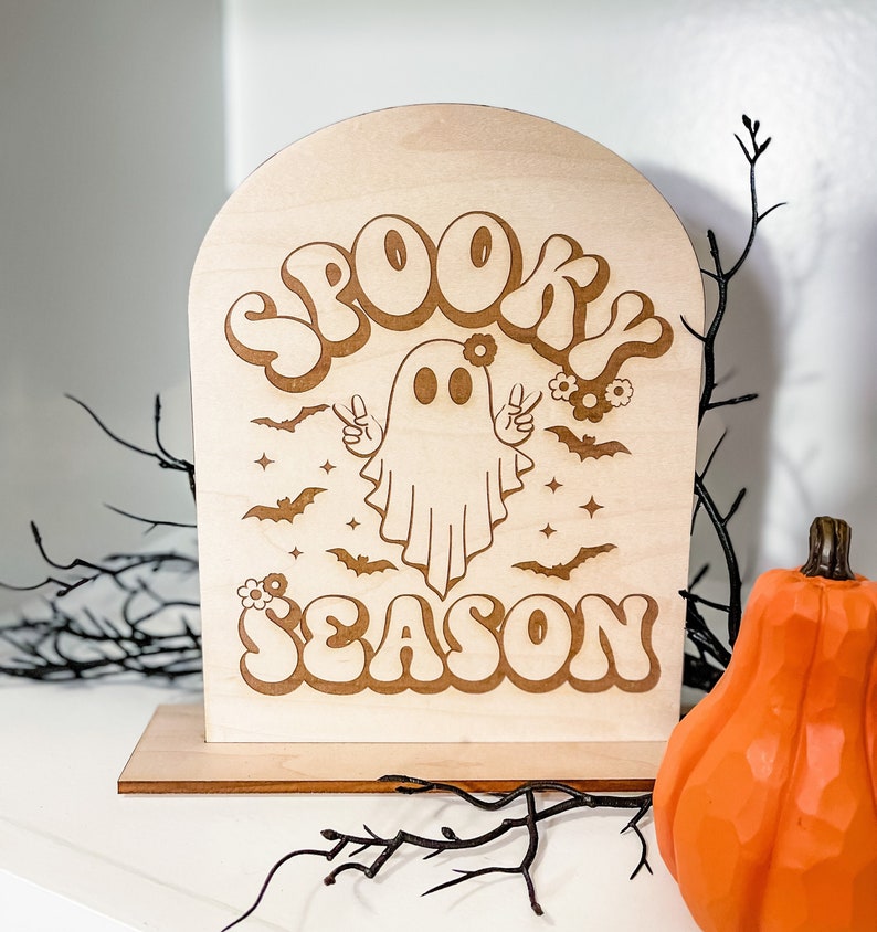 Może przedstawiać: Drewniany znak z duchem i tekstem "Spooky Season" w retro czcionce. Znak stoi na drewnianym stojaku i ma jasnobrązowy kolor.