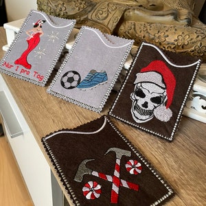 Puede incluir: Cuatro porta tarjetas de fieltro bordados diferentes. El primer porta tarjetas presenta una mujer roja con un vestido rojo con el texto "Nur 1 pro Tag". El segundo porta tarjetas presenta un balón de fútbol y un zapato azul. El tercer porta tarjetas presenta una calavera blanca con un gorro de Papá Noel rojo. El cuarto porta tarjetas presenta dos martillos cruzados y dos bastones de caramelo rojo y blanco.