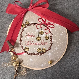 May include: A beige embroidery hoop with a red ribbon tied around it. The hoop features an embroidered wreath with the text "Frohe Weihnachten" and gold bells.