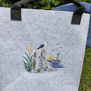 Könnte beinhalten: Eine hellgraue Filztasche mit schwarzen Henkeln und einer gestickten Küstenszene. Das Motiv zeigt eine Möwe auf einem Holzpfosten, ein Segelboot auf blauem Wasser, eine gelbe Sonne, hohes grünes Gras, einen Seestern und verschiedene Muscheln.