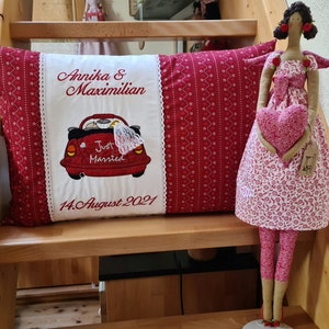 May include: A red and white embroidered pillow with the text "Annika & Maximilian" and "Just Married" with a red Volkswagen Beetle. The date "14. August 2021" is also embroidered on the pillow. A pink fabric doll with a heart is standing on a wooden step.
